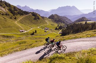 Drei Mountainbiker:innen auf Forsstraße mit Bergpanorama im Hintergrund